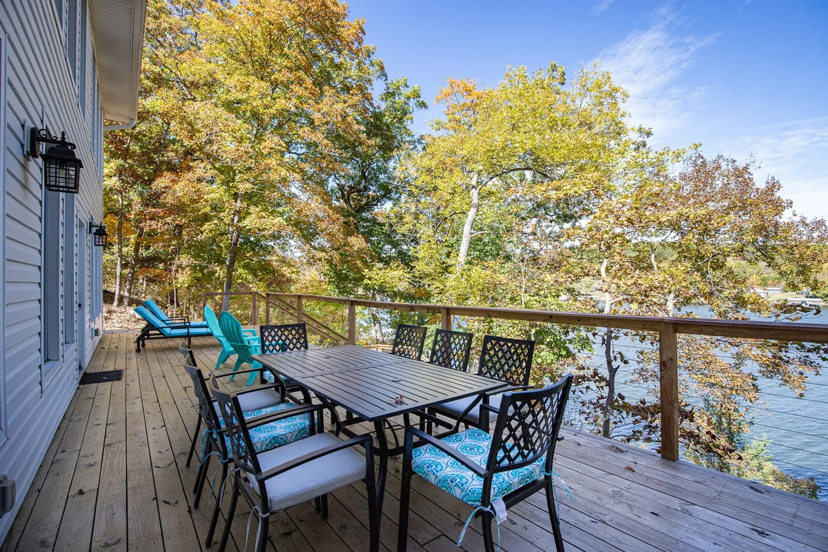 An exterior view featuring a spacious elevated deck surrounded by vibrant fall foliage, offering a dining table with seating and several outdoor chairs. The river is visible beyond the trees, creating a tranquil natural setting ideal for relaxation and family gatherings.