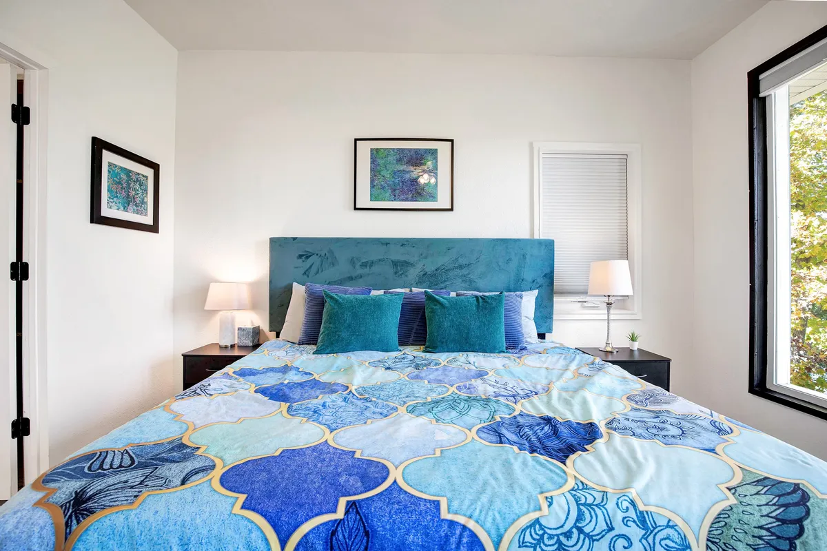 A cozy, relaxing bedroom with a bed topped by a patterned duvet and decorative pillows, complemented by artwork on the wall and nightstands with lamps on either side. The room is bright, airy, and designed for comfort and ease.