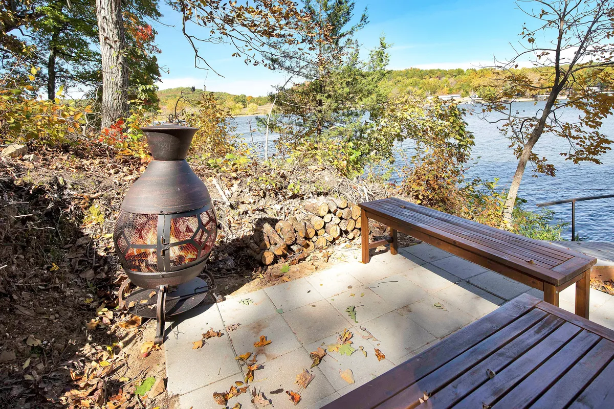 This image features a peaceful lakeside area with a chiminea (outdoor fireplace), a stack of firewood, a bench, and a clear view of the water. The scene is set for evening gatherings, storytelling, and roasting marshmallows as night falls over the lake.