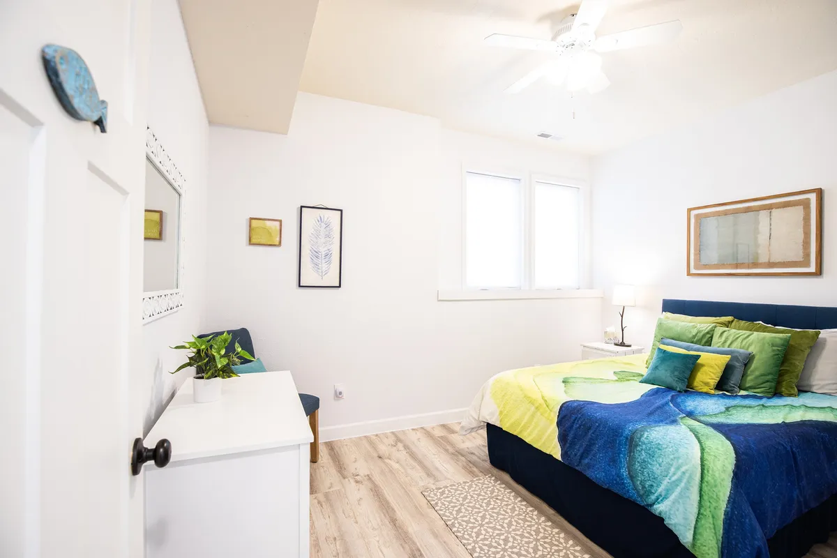 A cheerful bedroom furnished with a bed adorned with colorful bedding, flanked by two windows that let in ample natural light. The room also features decorative wall art, creating a lively yet inviting sleeping space.