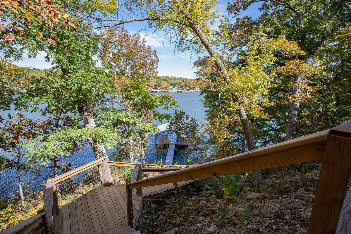 A wooden staircase leads down through lush green trees to a calm water surface and a dock, with inviting spaces for sitting and relaxing by the lake. The view highlights both access to the water and peaceful surroundings, ideal for fishing, swimming, or simply enjoying the outdoors.