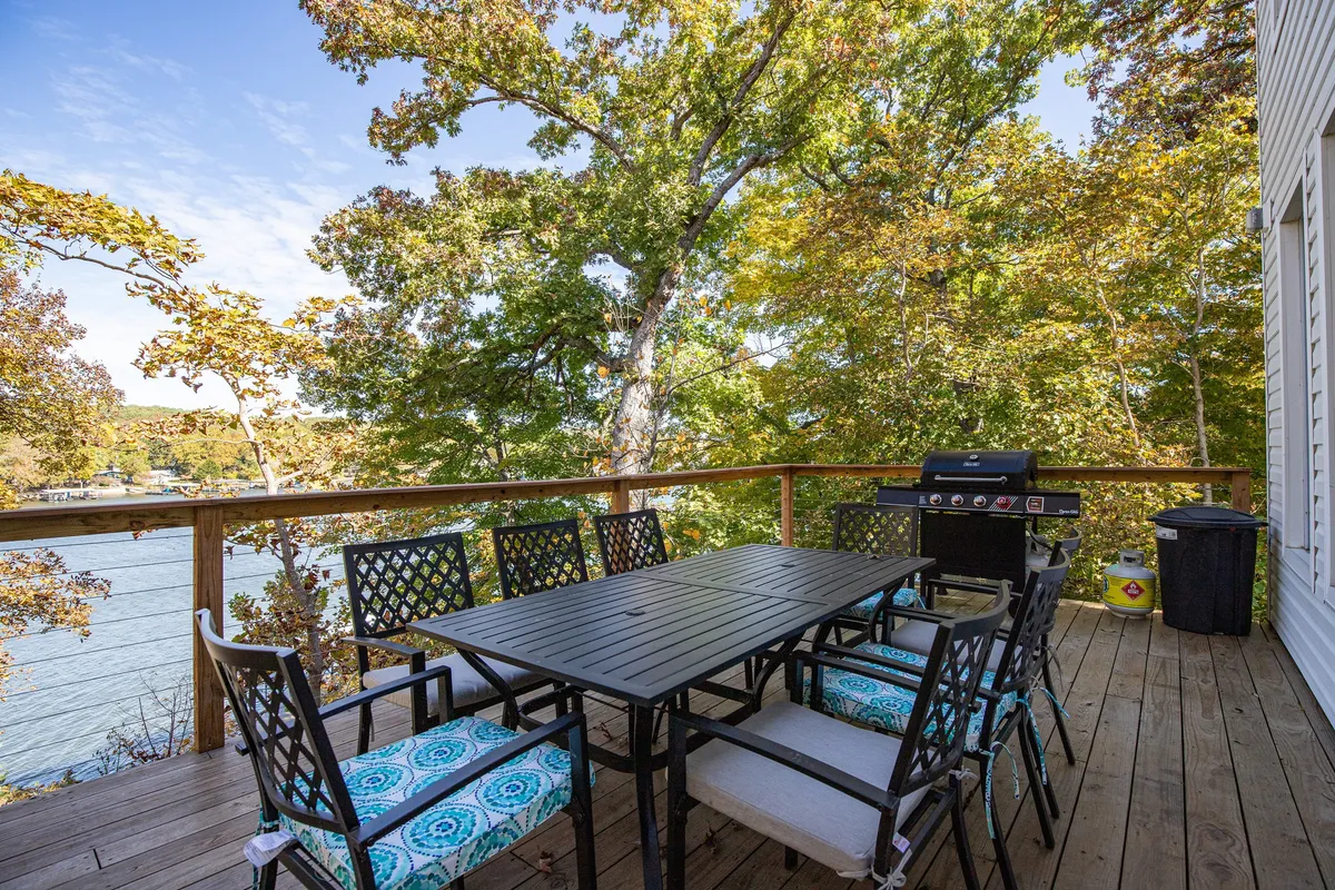An outdoor deck scene with an outdoor dining table, a grill, and sweeping lake views surrounded by lush trees. The mood is casual and serene, ideal for alfresco dining or sunset gatherings with friends and family.