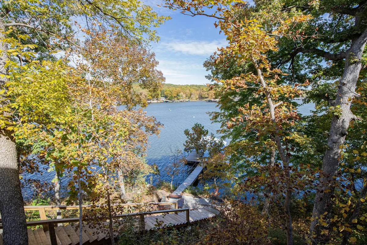 A tranquil lakeside scene featuring a wooden dock stretching out over the calm water, surrounded by lush trees and a winding pathway. The setting is perfect for boating, fishing, or simply relaxing by the water's edge. Autumn leaves dot the landscape, and the glassy surface of the lake reflects the soft morning sunlight.