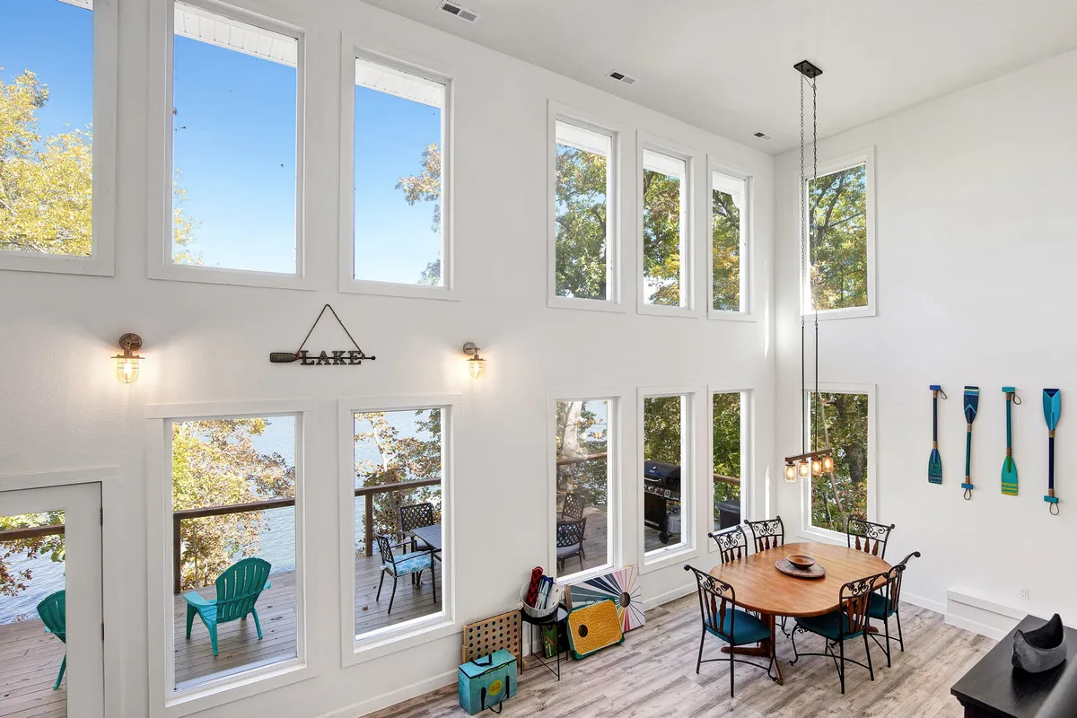 The image shows a spacious interior dining area with large windows offering panoramic views of the lake. A dining table is set for gatherings, and the space is accented with decorative paddles on the wall. The scene radiates warmth and tranquility, with natural light flooding in, inviting guests to relax, dine, and enjoy nature’s beauty from indoors.