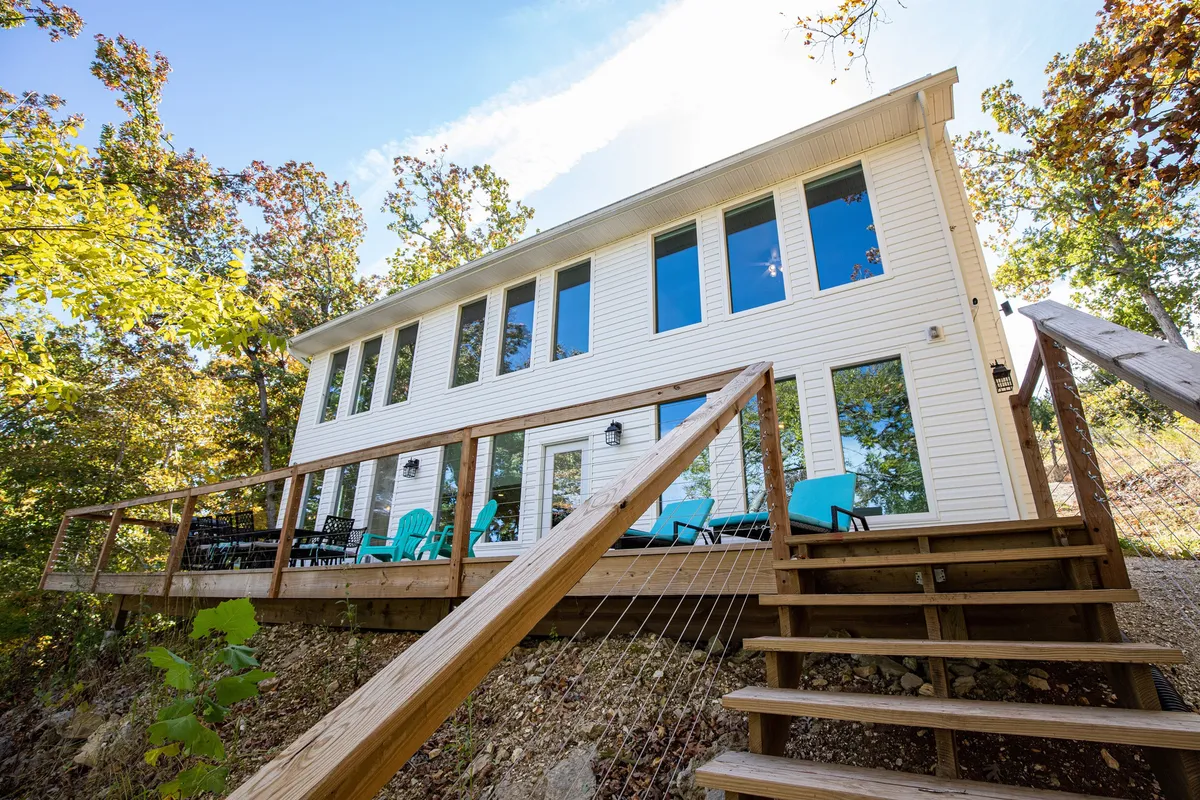 A wide deck with bright blue chairs faces out towards nature, attached to a house with large windows. The space is open and inviting, ideal for gatherings, relaxing, or enjoying meals outdoors while being close to the comforts of home.