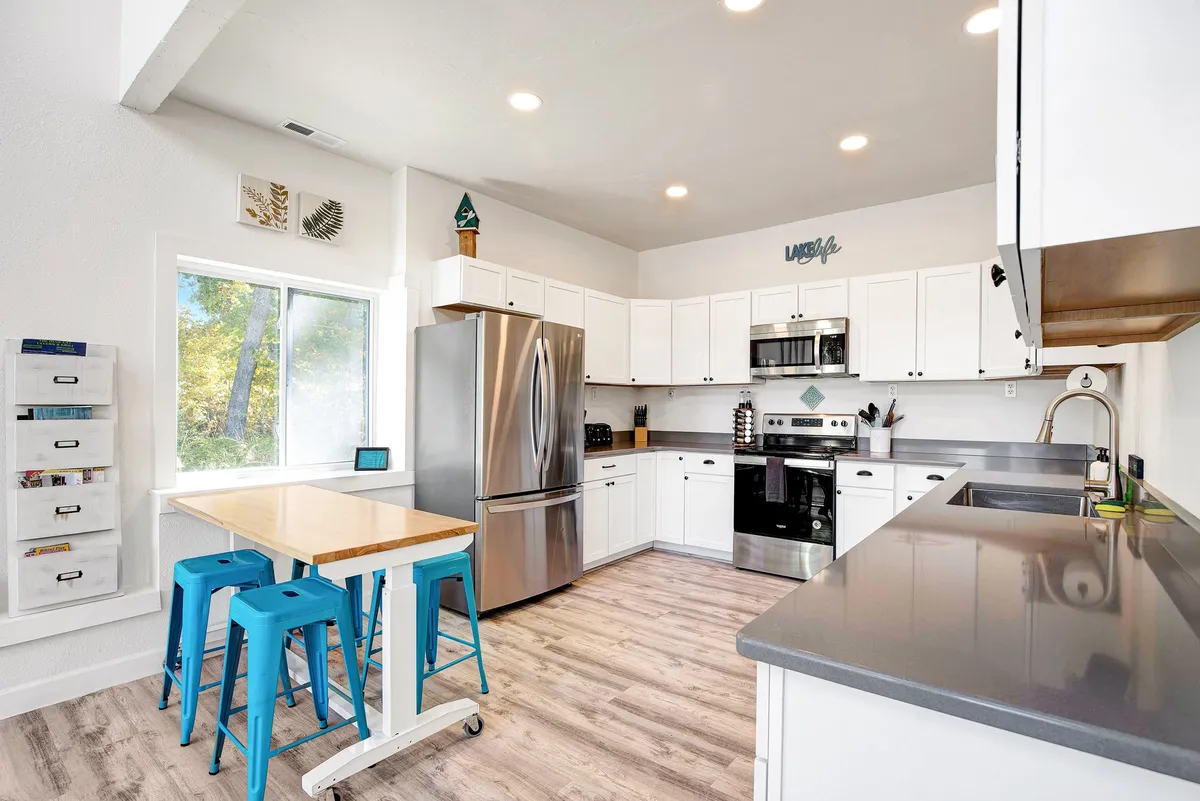A bright and cheerful kitchen and dining area featuring stainless steel appliances, a light wooden dining table, and blue bar stools. Natural light streams in through a large window, illuminating the space where cooking and casual gatherings take place.