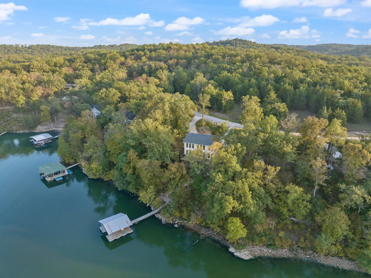 A broad exterior view captures the house nestled among trees, overlooking a serene lake with a dock stretching out into the water. The surrounding foliage and calm water set a peaceful tone, suggesting access to outdoor activities and exploration.