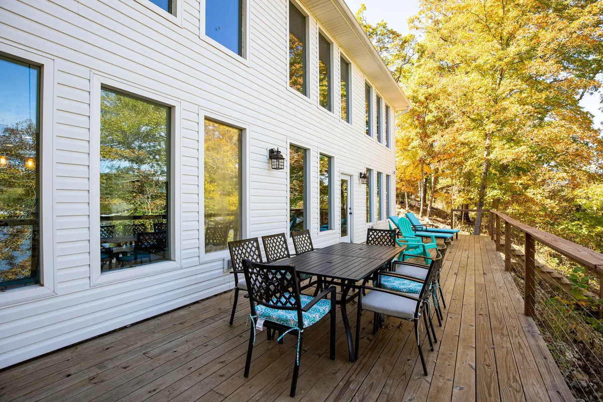 An inviting outdoor dining area on a deck, bordered by large windows and surrounded by trees. The table is set for gatherings, with colorful chairs beckoning guests to dine under the open sky. The setting suggests both relaxation and social connection, seamlessly bridging indoor comforts with the natural environment outside.